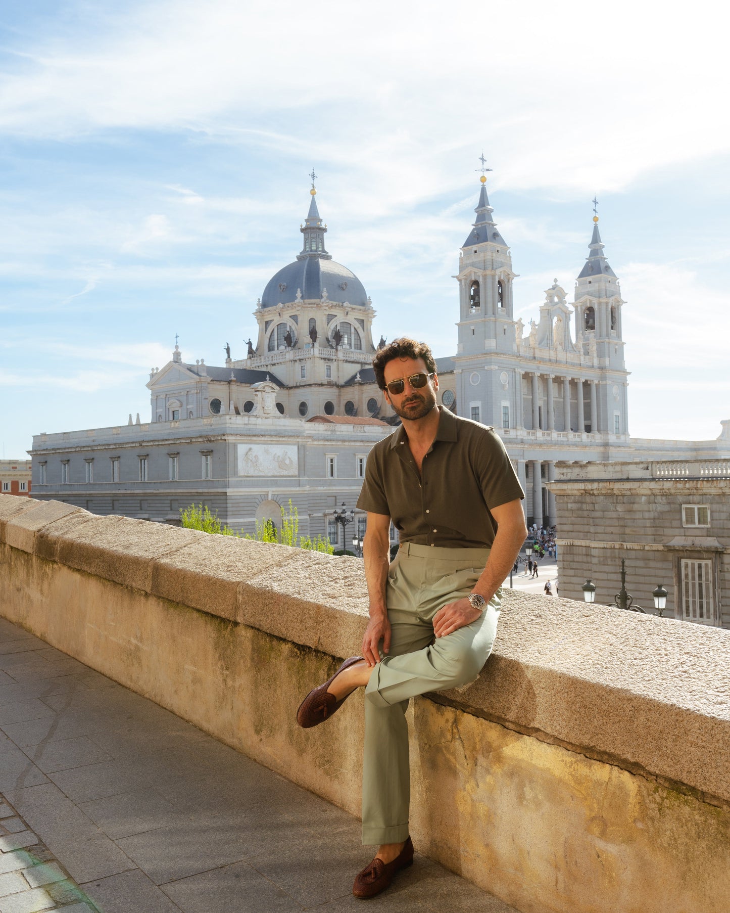 Model outside wearing mens custom polo cotton camp shirt by Luxire in olive green wearing sunglasses sitting on ledge